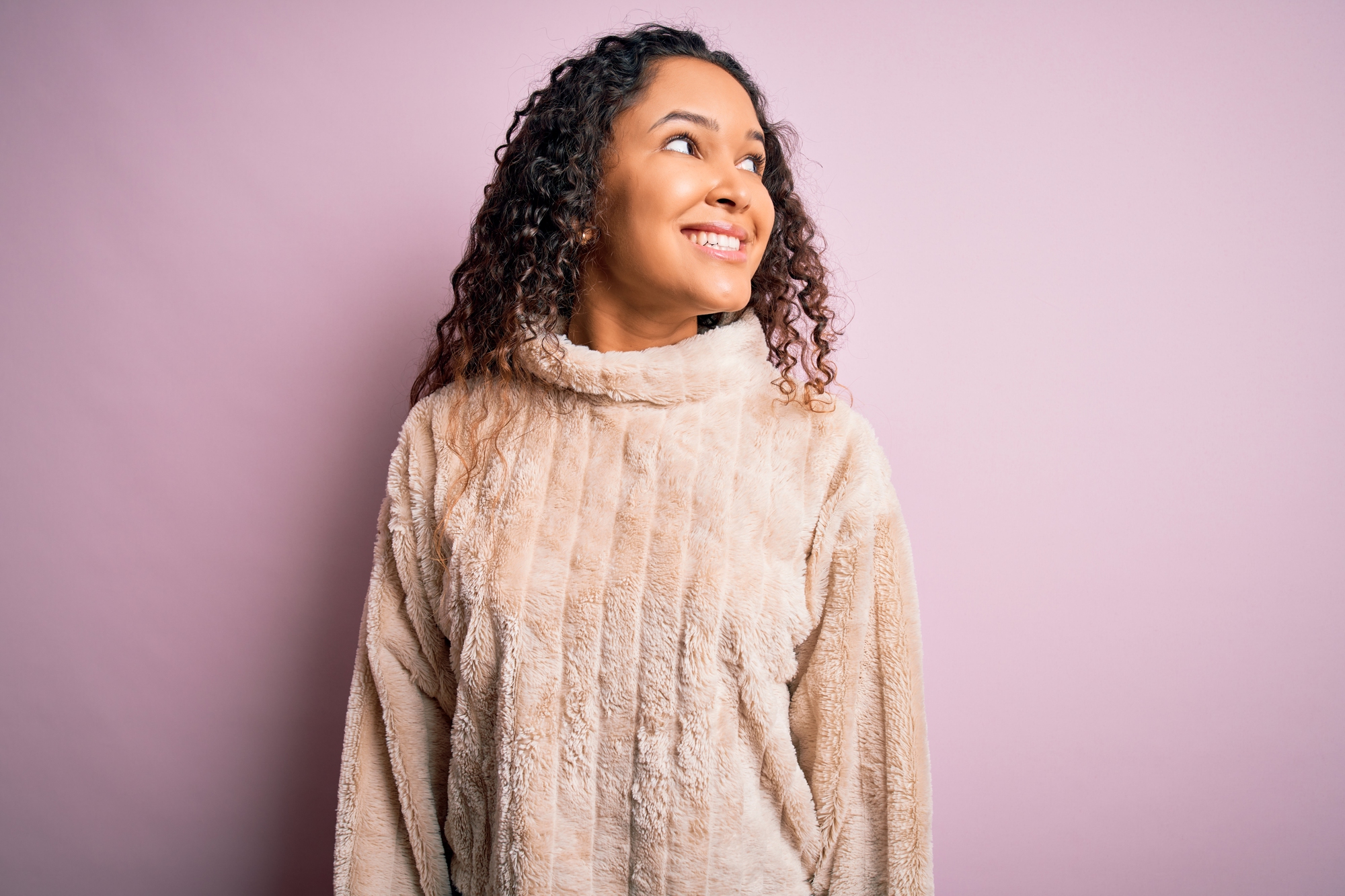 young woman with curly hair wearing casual sweater standing over pink background looking away to side with smile on face, natural expression. laughing confident.